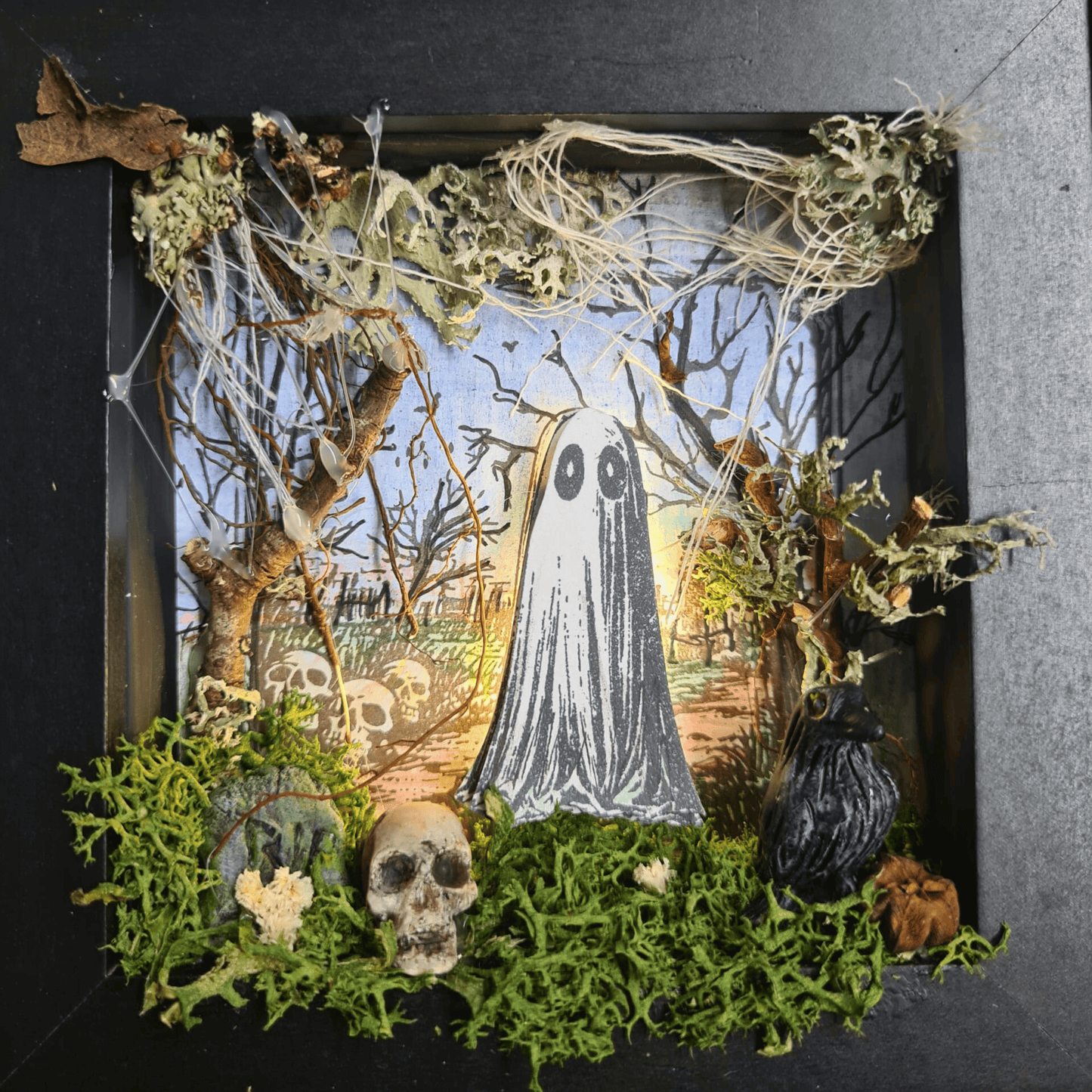 Completed 3D mixed media art scene from “The Little Haunting” kit, featuring a glowing ghost, sculpted tombstone, moss, fairy lights, and gothic frame.