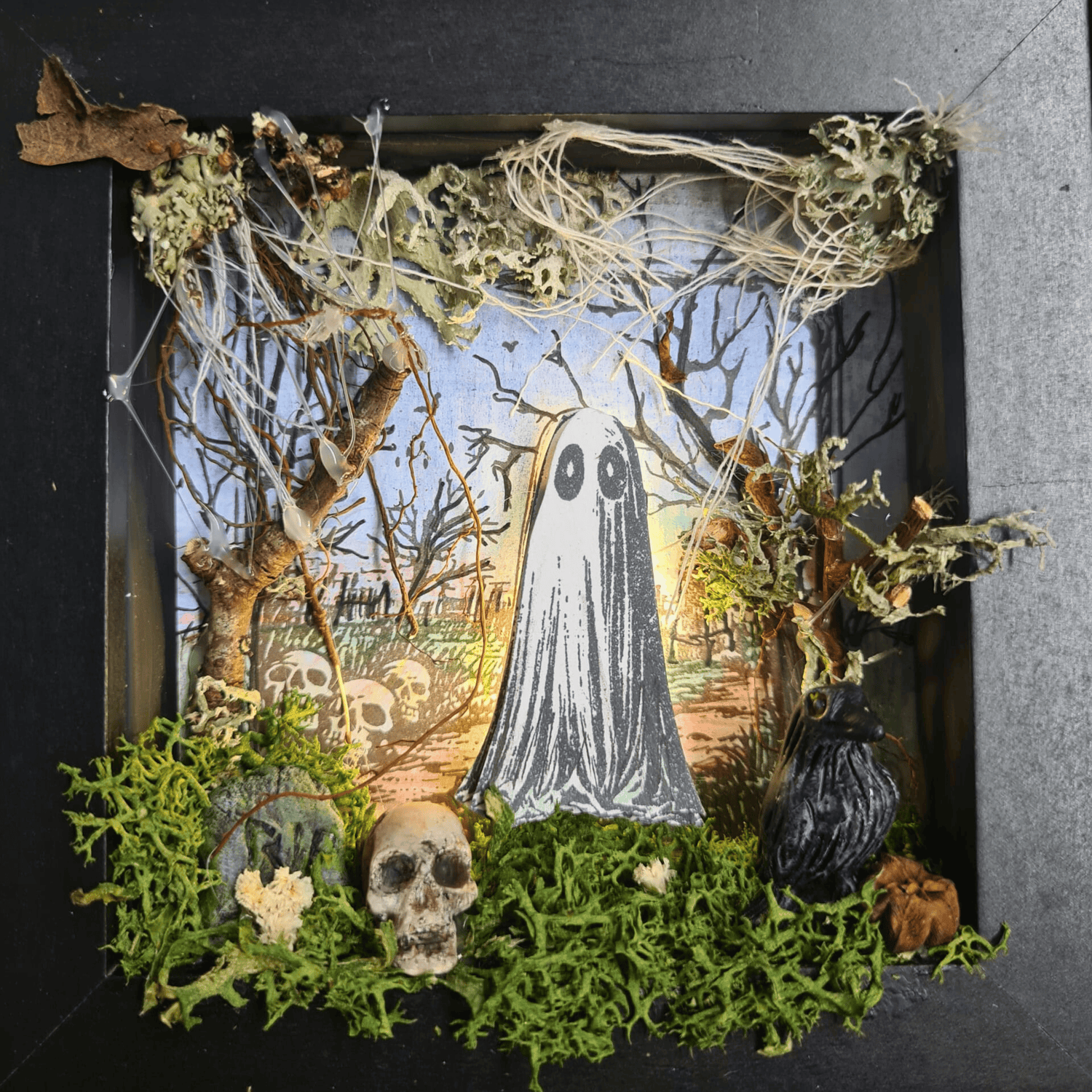 Completed 3D mixed media art scene from “The Little Haunting” kit, featuring a glowing ghost, sculpted tombstone, moss, fairy lights, and gothic frame.